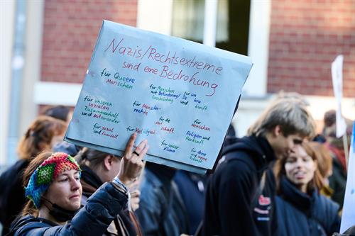 Person hält bei einer Demonstration ein handbeschriebenes Schild mit der Aufschrift „Nazis/Rechtsextreme sind eine Bedrohung“, umgeben von weiteren Teilnehmenden im Hintergrund.